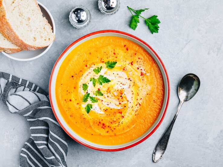 Ein Teller mit Zucchini-Möhren-Suppe in der Draufsicht. Daneben steht eine Schale mit Baguettescheiben, Gewürzstreuer und ein Löffel.