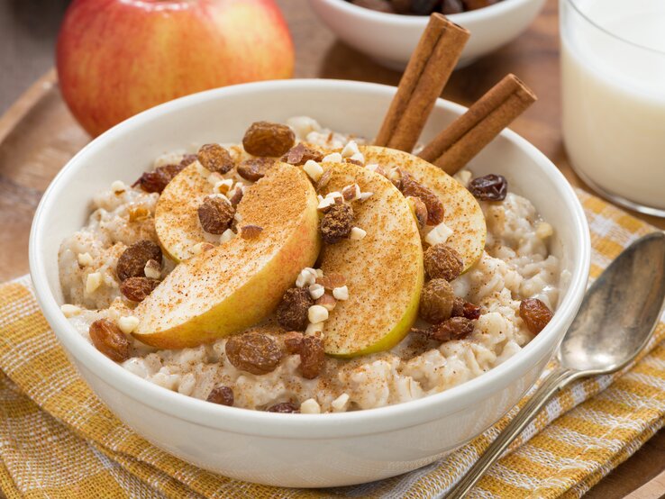 Eine weiße Schüssel mit Apfelstrudel-Porridge neben einem Löffel auf einem gelb-weiß-karierten Geschirrtuch.