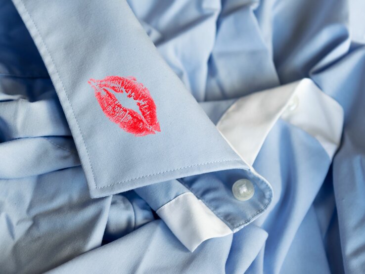 Auf der Manschette eines Herrenhemdes ist ein auffälliger roter Lippenstiftabdruck zu sehen. Der Abdruck hebt sich deutlich von dem hellen Stoff ab