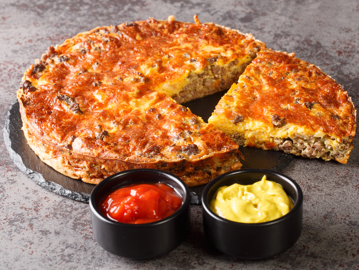 Cheeseburger-Kuchen auf Schieferplatte mit zwei Schälchen voll Senf und Ketchup.