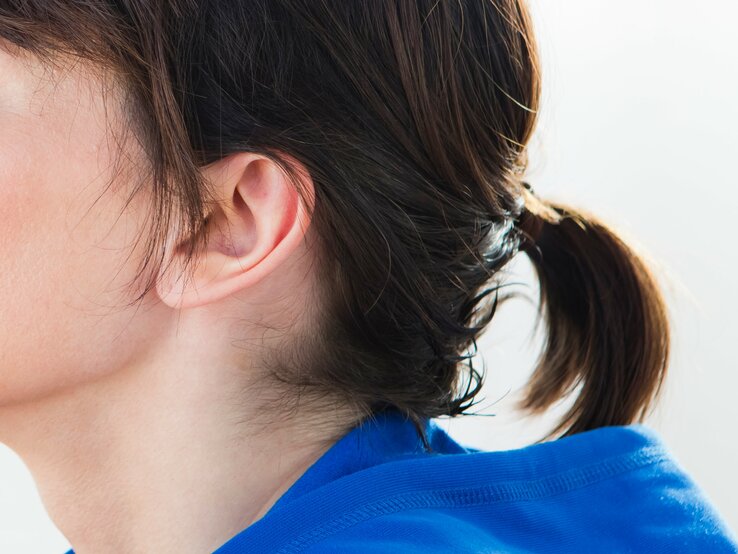 Teils eines weiblichen Kopfes mit Fokus auf dem Ohr und den Haaren. Das Ohr ist vollständig sichtbar und die Haare sind zu einem kleinen Pferdeschwanz gebunden, der mit einem unauffälligen Haargummi zusammengehalten wird.