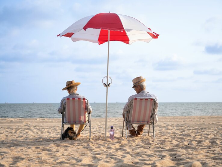 Zwei ältere Personen sitzen entspannt auf Strandstühlen unter einem rot-weißen Sonnenschirm am Strand. Sie tragen Strohhüte und sind dem Meer zugewandt, das sich im Hintergrund erstreckt. Daneben sitzt ein kleiner Hund im Sand.