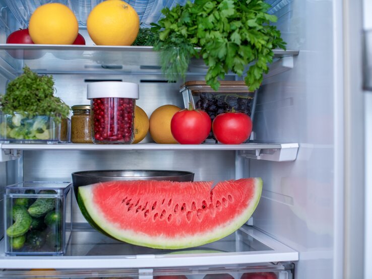 Ein geöffneter Kühlschrank in dem eine angeschnittene Wassermelone, Petersilie, Tomaten, Orangen und andere Lebensmittel gelagert werden.