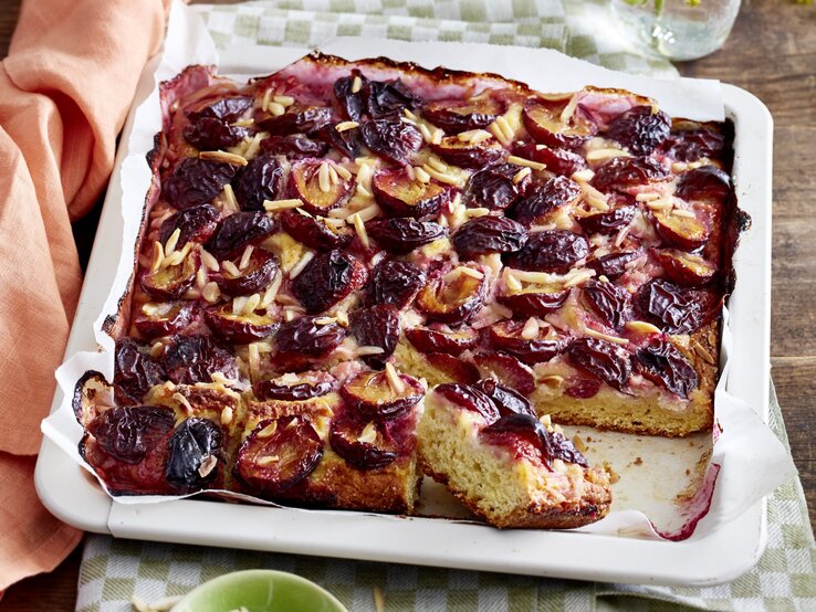 Angeschnittener Marzipan-Pflaumenkuchen in weißem Blech auf einem karierten Küchentuch, darunter Holz. 