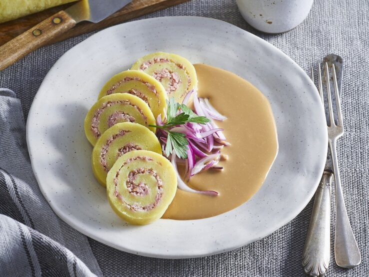 Kartoffelrouladen mit Schweinefleischfüllung auf Zwiebelsauce auf einem weißen Teller. Daneben liegt Besteck. 