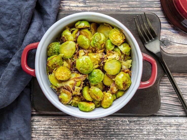 In der Draufsicht sieht man einen Topf mit Rosenkohl-Curry auf einem Brett, daneben eine Gabel und ein blaues Geschirrtuch.