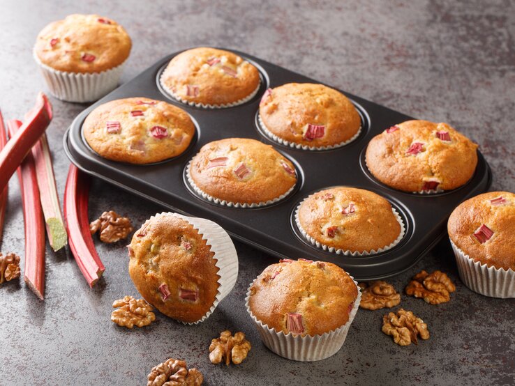 Rhabarber-Muffins mit Walnüssen in einer Muffinform, einige Muffins liegen daneben, außerdem frische Rhabarberstangen.