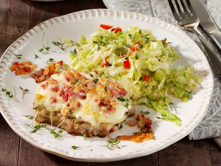 Ein weißer Teller auf einem dunklem Holztisch mit einer grau-braunen Tischdecke. Auf dem Teller ist etwas Krautsalat und das gratinierte Stück Fleisch mit Speck und Käse.