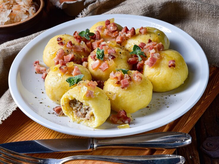 Die Knödel auf einem weißen Teller. Dieser steht wiederum auf einem Holzschneidebrett mit Messer und Gabel.