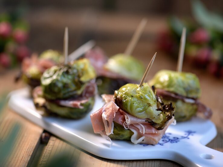 Rosenkohl-Spießchen mit Salami und Prosciutto auf einem weißen Brett mit unscharfem Hintergrund.