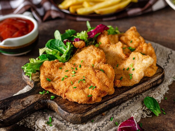 Ein Holzbrett mit drei Schnitzeln und etwas Schnittlauch auf einem dunklen Holztisch. Auf dem Tisch sind auch unscharf Pommes, Ketchup und Salat zu erkennen.