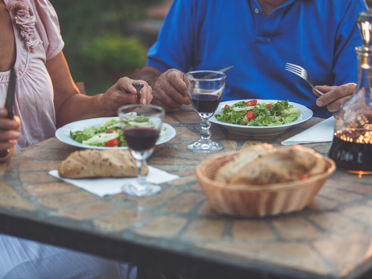 Zwei ältere Personen essen Salat und Brot an einem rustikalen Tisch im Freien, mit Rotweingläsern und einer Karaffe im Abendlicht.