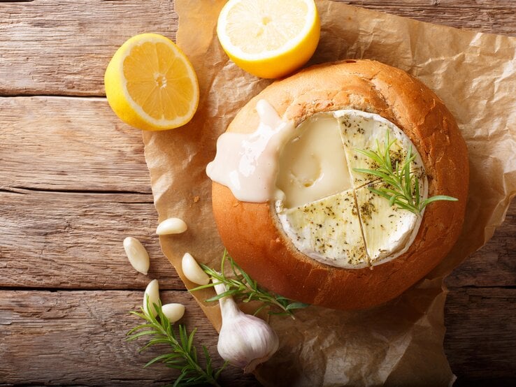 Auf einem hölzernem Untergrund liegt ein Packpapier. Darauf liegt ein ausgehöhlter Brotlaib. In dem Brotlaib ist ein Camembert, auf dem ein Zweig Rosmarin zu sehen. Neben dem Brot liegen eine Knoblauchknolle, Knoblauchzehen und ein weiterer Rosmarinzweig.