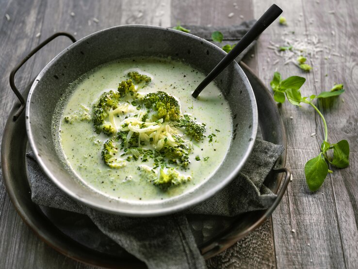 Auf einem rustikalen Holztisch sehe ich eine eiseren Pfanne in der eine schöne Schüssel aus Metall steht. In der Schüssel ist eine leuchtend grüne, cremig-pürierte Suppe, die mit ganzem Brokkoliröschen garniert ist. Die Suppe sieht sehr lecker aus! neben dem Ensemble liegt ein Zweig frisches Basilikum.