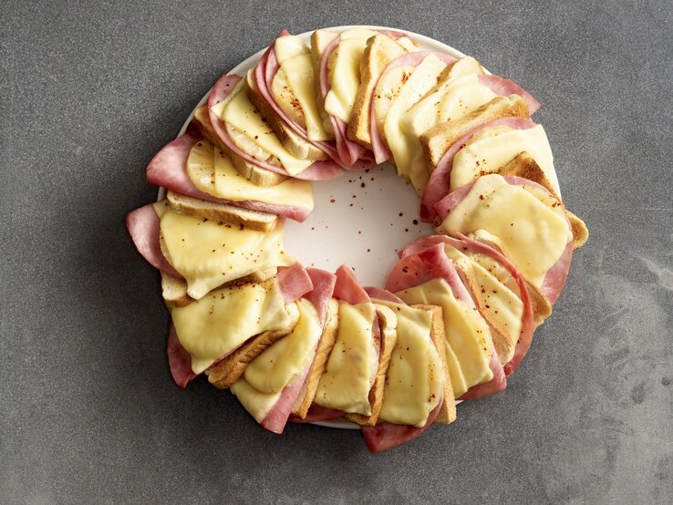 Hawaii-Toastbrot-Ring, bestehend aus abwechselnden Lagen von Gouda, Schinken, Ananas und Toastscheiben. In einem Ring aneinandergereit überbacken und mit Chiliflocken bestreut auf einem weißen Teller serviert. 