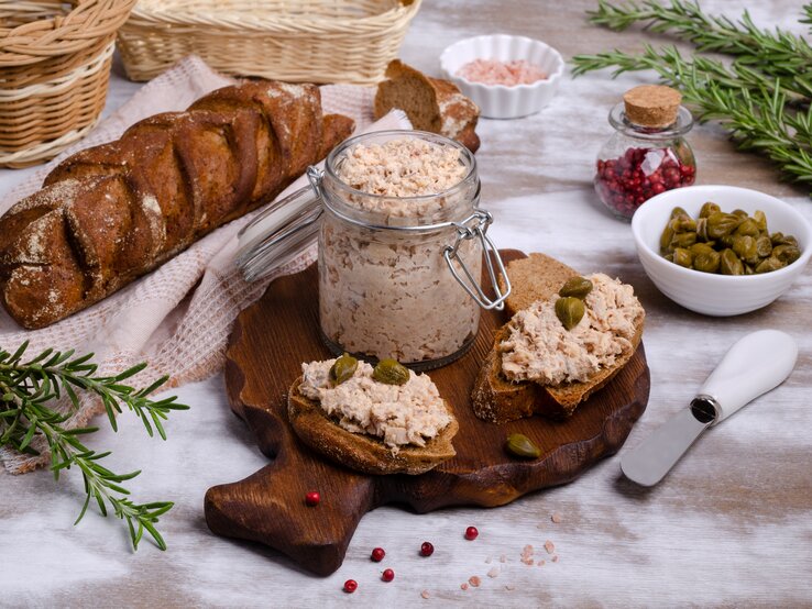 Frontal: Ein gedeckter Tisch mit Tischdecke, auf dem ein Glas Lachs-Rillette , eine Schale mit Kapern, ein Glas mit rosa Salz, ein Brotkorb, frischer Rosmarin und ein Brot liegen.