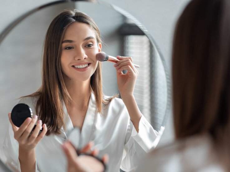 Junge Frau schminkt sich vor einem runden Spiegel. Sie hält einen Make-up-Pinsel und ein Kompaktpuder in ihren Händen und lächelt dabei leicht. Die Frau trägt ein weißes, locker sitzendes Hemd und hat glattes, braunes Haar. 