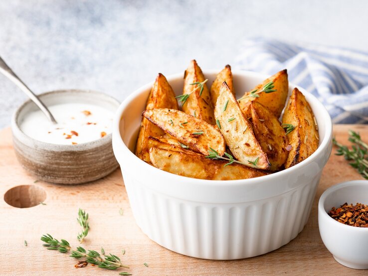 Kohlrabi-Wedges mit Thymian in einer weißen Schale auf einem Brett, darauf außerdem eine Schale Dip und eine Schale mit getrockneten Gewürzen. 