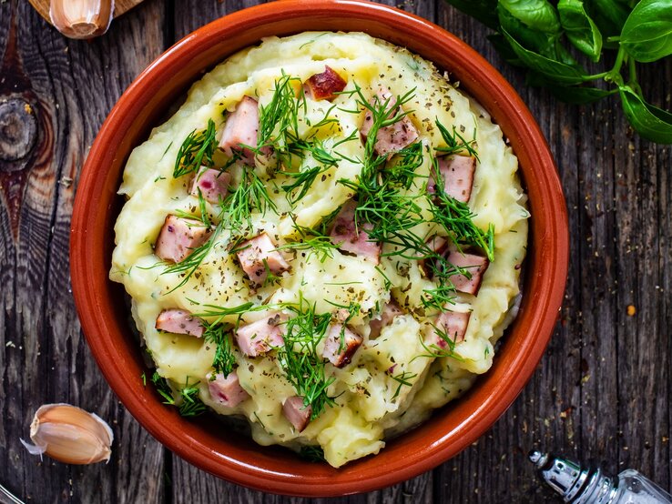 Dill-Kartoffelpüree mit Leberkäse in roter Schüssel auf dunklem Holz. 