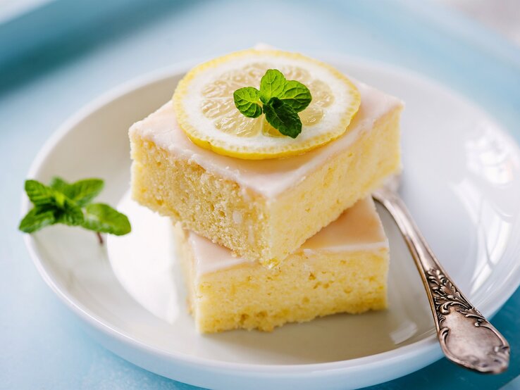 Zwei Stück Selterskuchen mit Zuckerguss und einer Zitronenscheibe und Minze garniert auf einem Teller mit einer Kuchengabel.