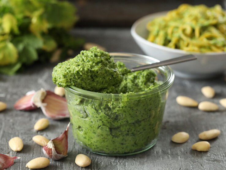 Frontal: eine Glas selbstgemachtes Mandel-Petersilien-Pesto steht auf einem dunklen Untergrund aus Holt. Drum herum liegen Mandeln ohne Haut und ungeschälte Knoblauczehen.