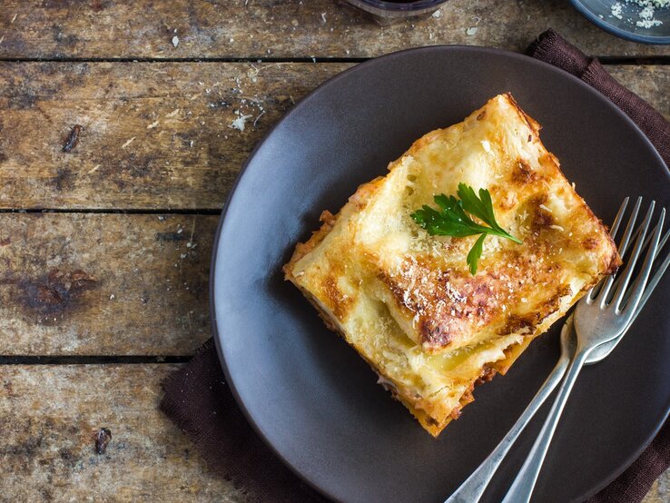 Ein Stück Lasagne auf einem rustikalen Holztisch von oben fotografiert. Darüber eine kleine Schale mit geriebenem Käse und ein Glas mit Rotwein.