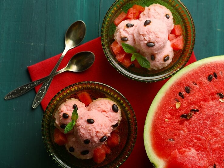 Draufsicht: Dunkelblauer Untergrund, zwei Schälchen aus Glas mit selbst gemachten Wassermelonen-Eis, dass auf einem bett von wassermelonen-Würfeln liegt und mit Wassermelonen-Kernen und einem Minzblatt dekoriert ist. Daneben liegt eine aufgeschnittene Wassermelonen-Hälfte.