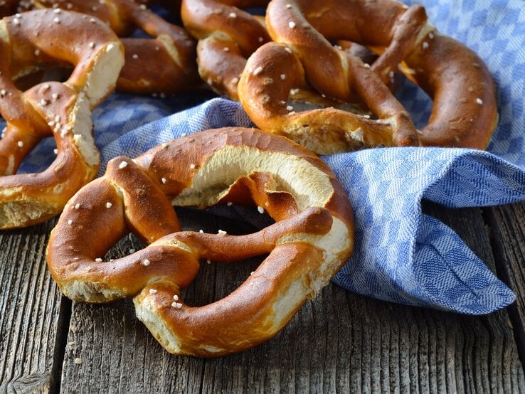 Draufsicht: Auf einem Holutisch liegen frische bayerische Brezen azf einem groben weiß-blau kariertem Tuch.