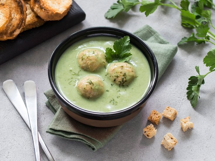 Zucchinisuppe mit Hackfleisch-Bällchen in schwarzer Schüssel auf hellem Untergrund. Daneben Kräuter, Löffel und geröstetes Brot. 