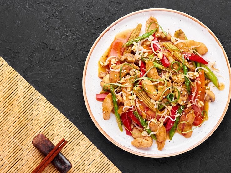 Ein weißer Teller mit dem Chop Suey auf dunklem Untergrund mit Stäbchen und Bambusmatte unten rechts von oben fotografiert.  