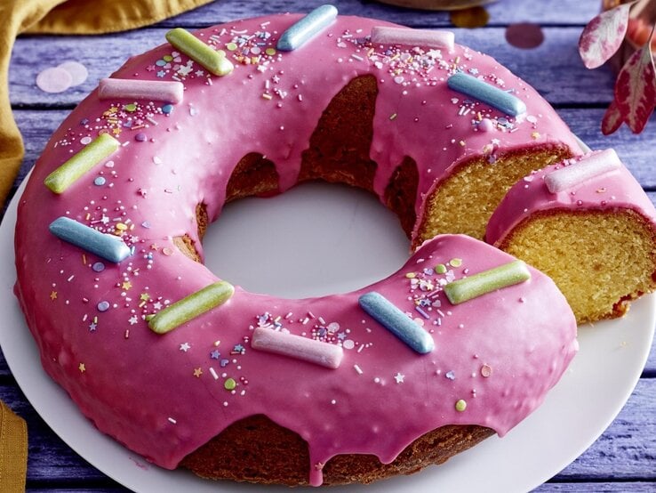 Auf einem Teller liegt ein  Donut-Kuchen in Kranzform. Er ist mit Glasur überzogen und mit bunten Streuseln und Süßigkeiten dekoriert.