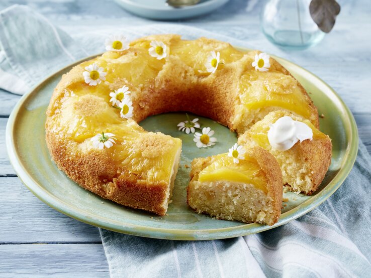 Auf einem Porzellanteller liegt ein Ananas-Kokos-Kuchen in Kranzform. Er ist mit weißen essbaren Blüten dekoriert. 