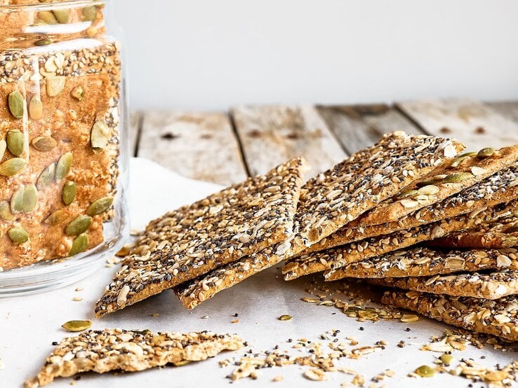 Mehrere Knäckebrote auf einem Holztisch mit Backpapier und einem Glas mit dem Knäckebrot.