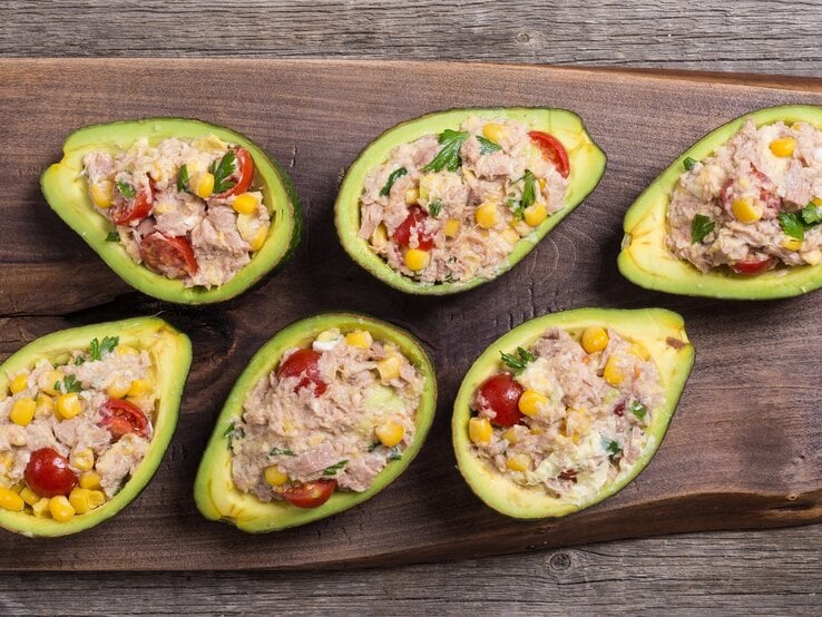 Ein Holzbrett mit sechs Avocadohälften und dem Thunfischsalat. Alles auf einem rustikalem Holztisch direkt von oben fotografiert.