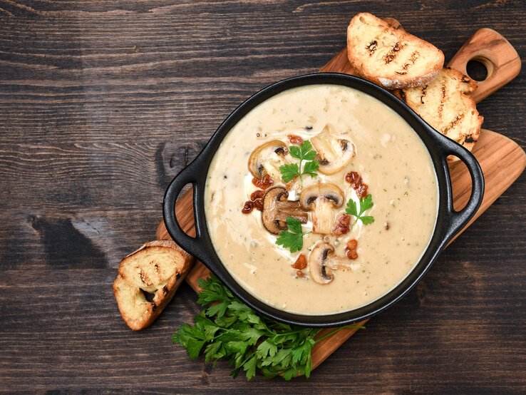 Draufsicht: Ein Holzbrettchen auf dem einen schwarze Schüssel steht. In der Schale ist eine alpenländische Kümmelrahmsuppe mit Pilzen. Drum herum liegen drei Scheiben geröstetes Weißbrot und frische Petersilie.