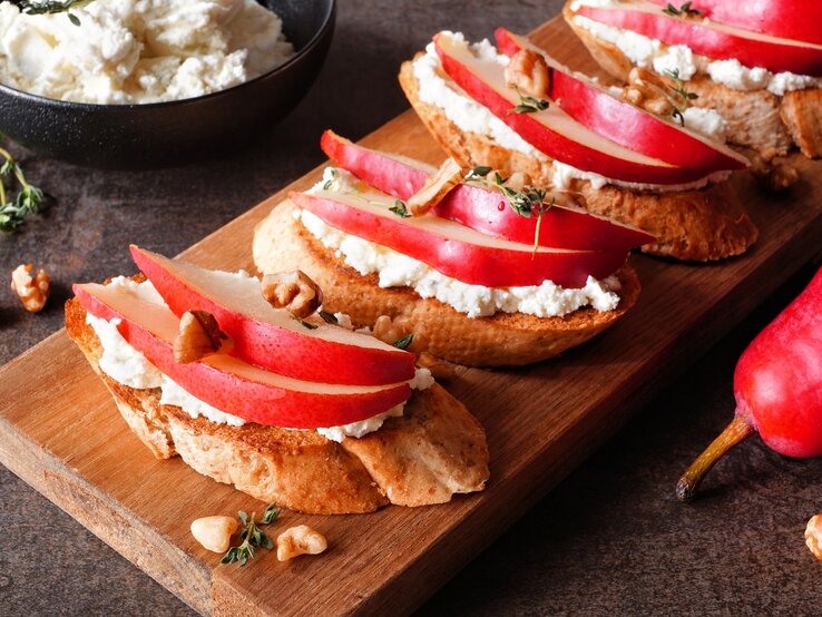 Draufsicht schräg: Ein Holzbrettchen mit vier Hüttenkäse-Sandwich mit Birne und Walnüssen liegt auf einem dunklen Holzuntergrund. Im Hintergrund ist eine dunkle Schüssel mit Hüttenkäse. Neben dem Brettchen ist ein Teil einer roten Birne zu sehen.