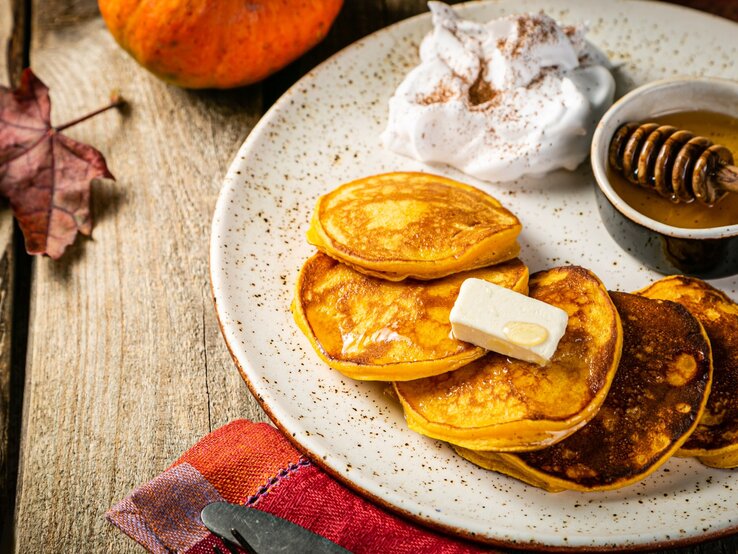 Vier Kürbis Pancakes auf einem hellen Teller mit Honig und etwas geschlagener Sahne. Alles von schräg oben auf einem rustikalen Holztisch mit herbstlicher Deko fotografiert.