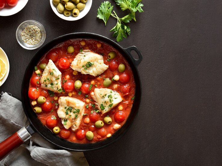 Draufsicht: Eine mediterrane Fischfilet-Pfanne auf dunklem Untergrund. Drum herum liegen Zitronenscheiben, Petersilie und Gewürze.