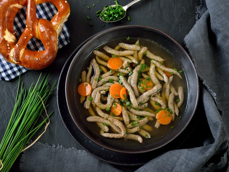 Draufsicht: Dunkler Untergrund, dunkles Tuch, daneben ein Teller bayerische Rindersuppe mit Leberspätzle. Daneben liegen Bund Schnittlauch, Schnittlauchröllchen und eine Breze auf einem kariertem Tuch.