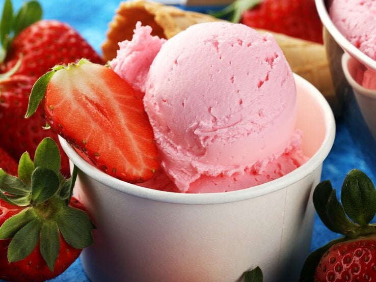 Der Erdbeer-Eisbecher zwischen vielen Erdbeeren und einer Waffel auf einem blauen Tisch.