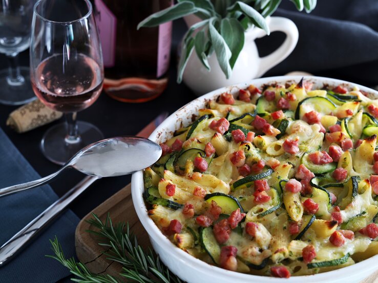 Würziger Zucchini-Nudelauflauf mit saftigen Speckwürfeln bestreut und überbacken mit Käse, serviert in einer runden, weißen Auflaufform. Daneben stehen Gläser und ein wenig Dekoration. 