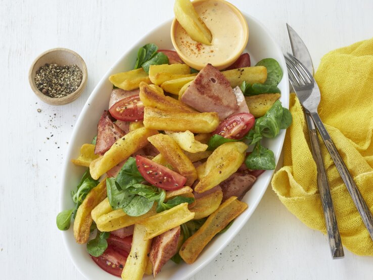 Draufsicht: Heller Untergrund auf dem ein ovaler Teller mit bayerischen Pommes mit Leberkäse und Obazda-Dip steht. Garniert mit ein paar Blättern Feldsalat und Tomatenspalten. Rechts daneben eine gelbe Stoffservierte und rustikales Holzbesteck.