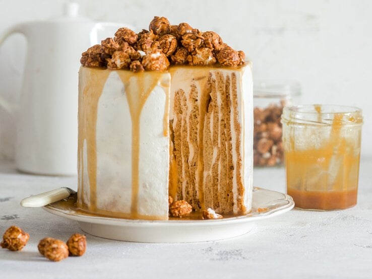 Angeschnittener Nusskuchen mit Karamell und Karamellpopcorn darauf, daneben Glas mit Karamell, heller Hintergrund. 