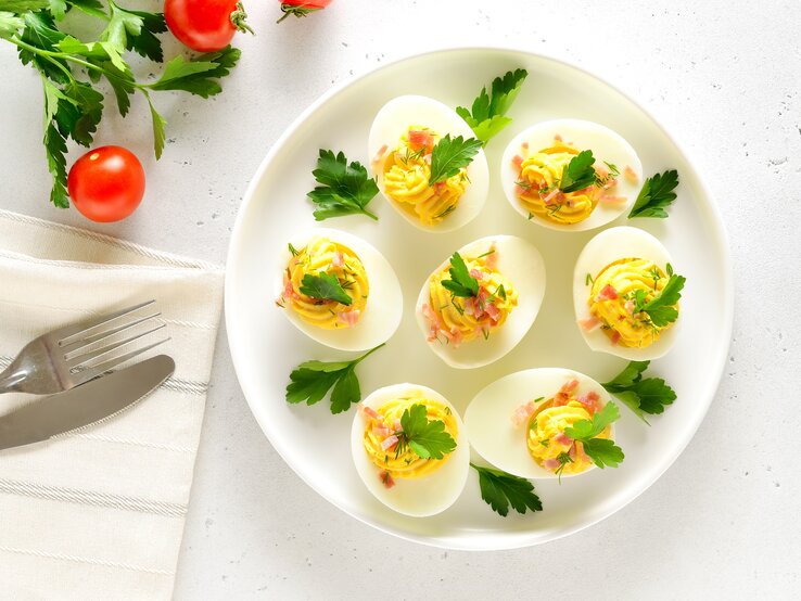 Draufsicht: Ein weißer Untergrund. Gefüllte Eier mit Speck auf einem weißen Teller. Daneben sind Tomaten, frische Petersilie und ein Besteck.