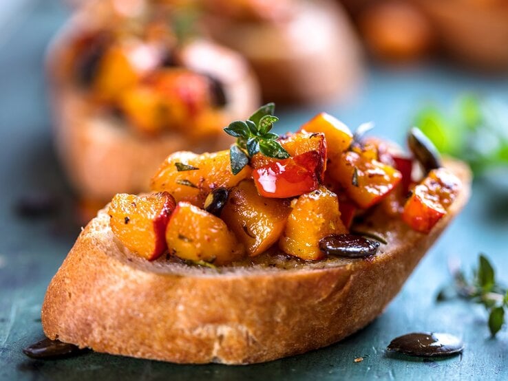 Eine Kürbis-Bruschetta mit Kürbiskernen und Kräutern auf einem Stück Baguette, im Hintergrund unscharf weitere Bruschettas.