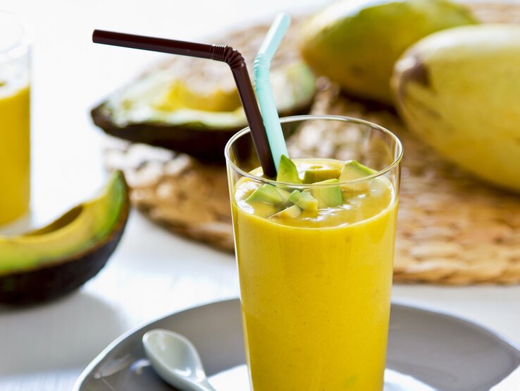 Ein Glas mit dem gelben Smoothie auf einem hellen Tisch mit Avocado und Mango im Hintergrund.