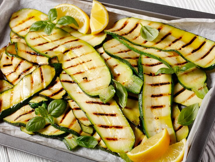 Auf einem mit Backpapier ausgekleideten Backblech liegen Zucchini, die gegrillt worden sind. Auf und zwischen ihnen liegen Zirtonenschnitzer und Basilikumblätter.