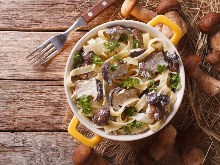 Draufsicht: Auf einem Holzboden steht ein Topf mit Tagliatelle mit Steinpilzen. Drum herum liegen eine Gabel und frische Steinpilze.