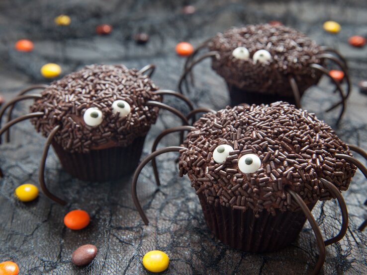 Süße Spinnen-Muffins mit Halloween auf dunklem Untergrund. Daneben liegen Schokolinsen in verschiedenen Farben. 