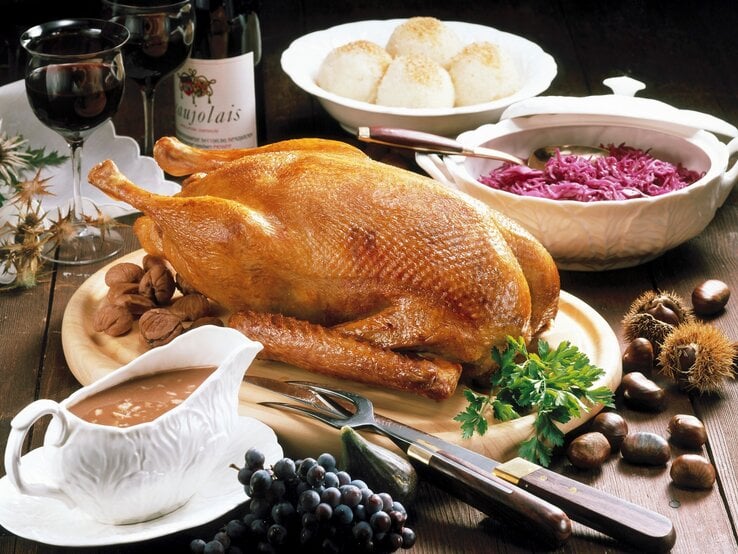 Gefüllter Gänsebraten mit Apfel und Marone auf Festtafel mit Beilagen. Im Hintergrund eine Flasche Rotwein und zwei Gläser und Dekoration. Draufsicht.